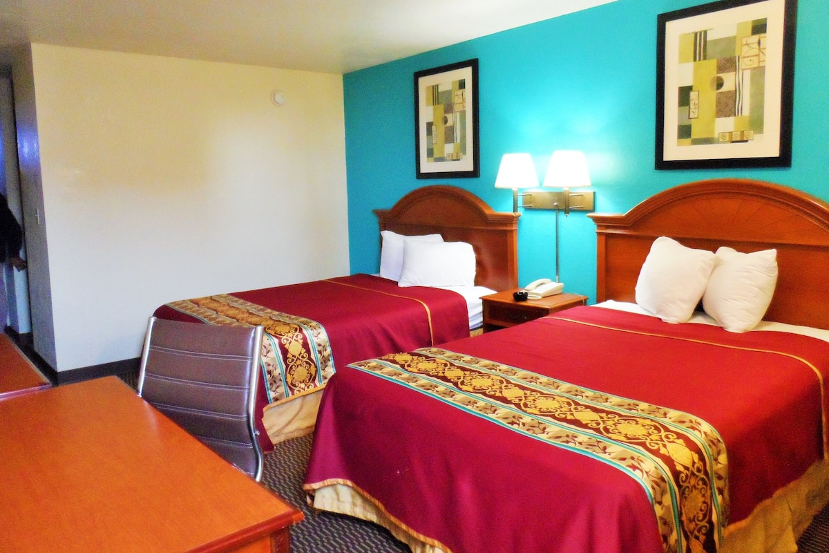 A hotel room with two beds, a desk, and a chair. There are two bedside tables with lamps and a telephone. The walls are painted in two colors, and there are two framed pictures on the wall.