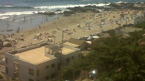 A beach with people and umbrellas, a rocky shoreline, and buildings in the background.