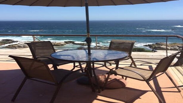 A round table with four chairs and a large umbrella, overlooking a rocky coastline and the ocean.