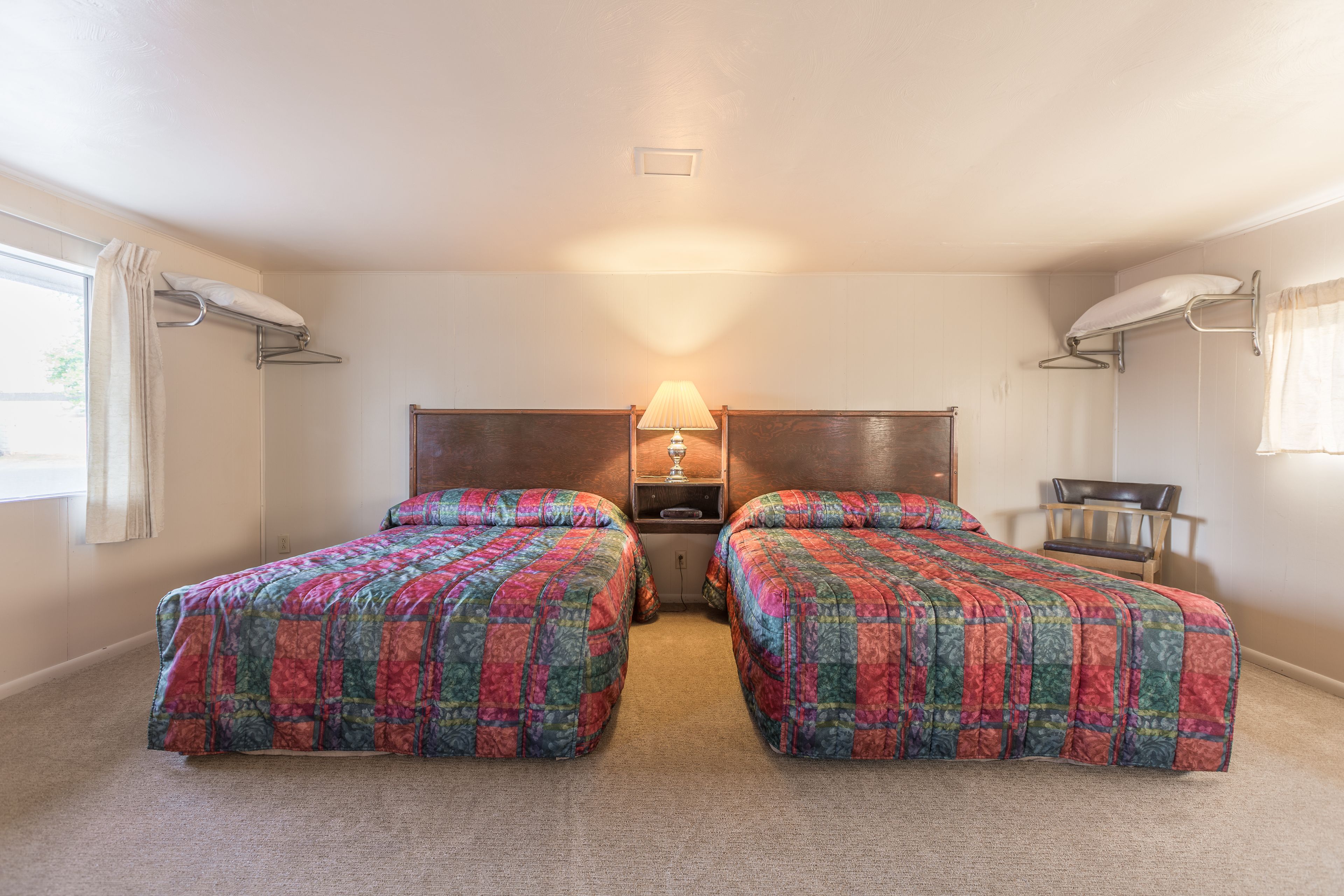 Una habitación con dos camas, una mesita de noche con lámpara, una silla y una ventana con cortinas.