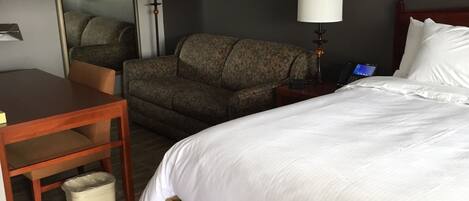 A hotel room with a bed, a desk, a chair, a trash bin, and a lamp.