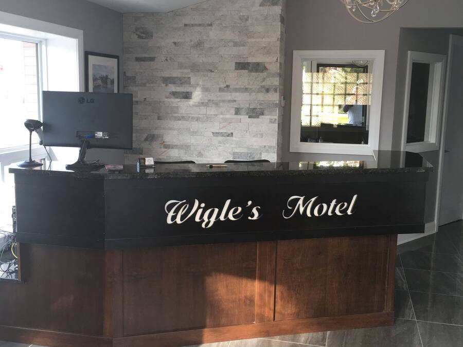 Een receptiebalie met de tekst 'Wigle's Motel', op de achtergrond een stenen muur, een kroonluchter en een groot raam met blinds.