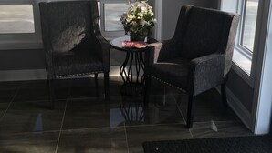 Une pièce avec deux fauteuils sombres, une petite table ronde ornée d’un vase de fleurs et une vue sur un parking, visible à travers de grandes fenêtres.