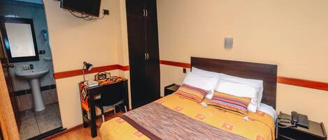 A hotel room with a bed, bedside table, desk, chair, wardrobe, and a television mounted on the wall.