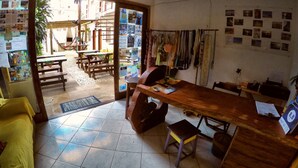 A room with a wooden table, a yellow couch, and a view of an outdoor area with a hammock and a tiled floor. - Buddy's Hostel e Pousada (Itacaré)