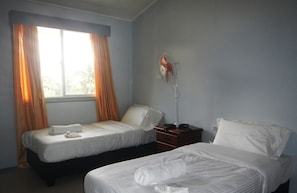 Two single beds with white mattresses and pillows, a standing fan, and a nightstand with a lamp.