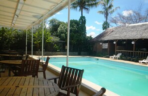 Relax by the pool surrounded by lush greenery and a thatched-roof building.
