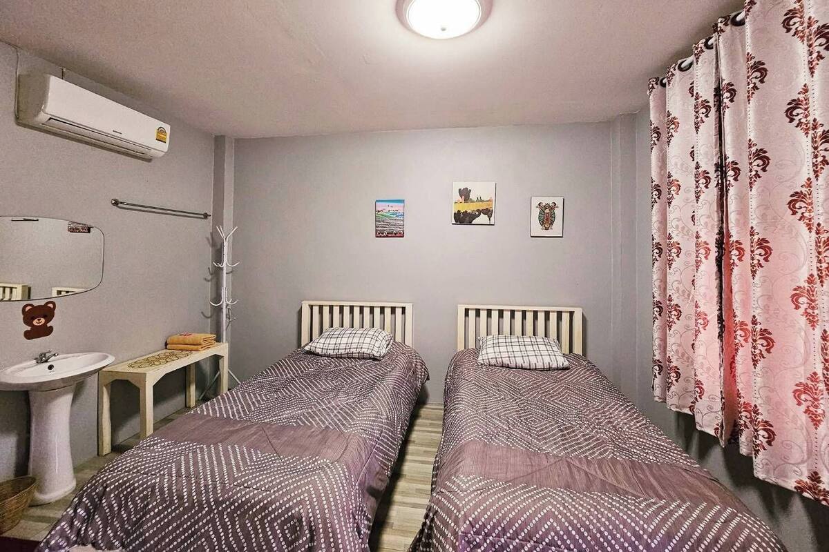 A small, well-lit bedroom with two single beds, a bedside table, a mirror, a wall-mounted air conditioner, and a window with curtains.
