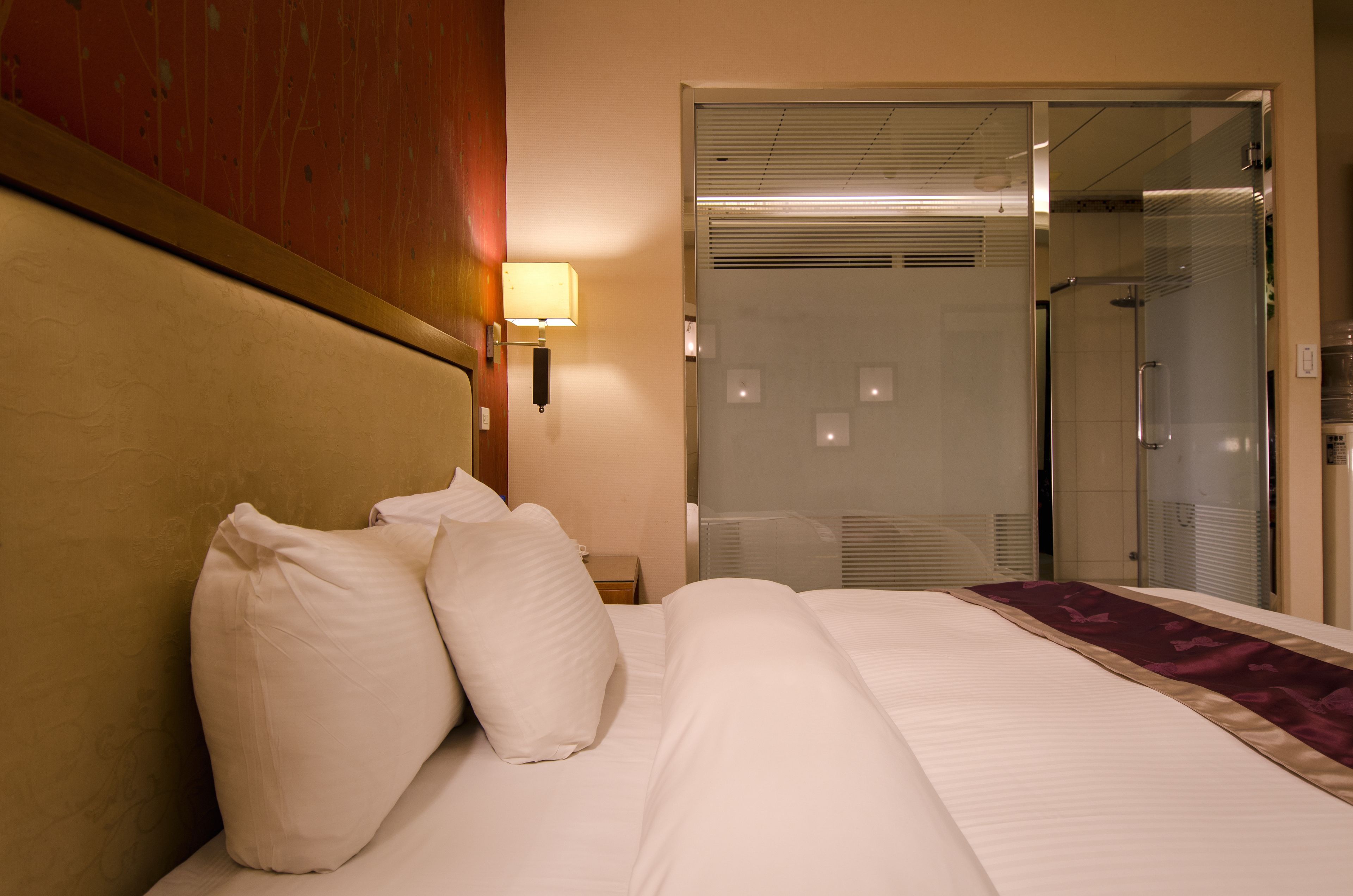 Une chambre d’hôtel comprenant un lit, une table de chevet, une lampe, une porte coulissante donnant sur une cabine de douche et une tête de lit en bois.