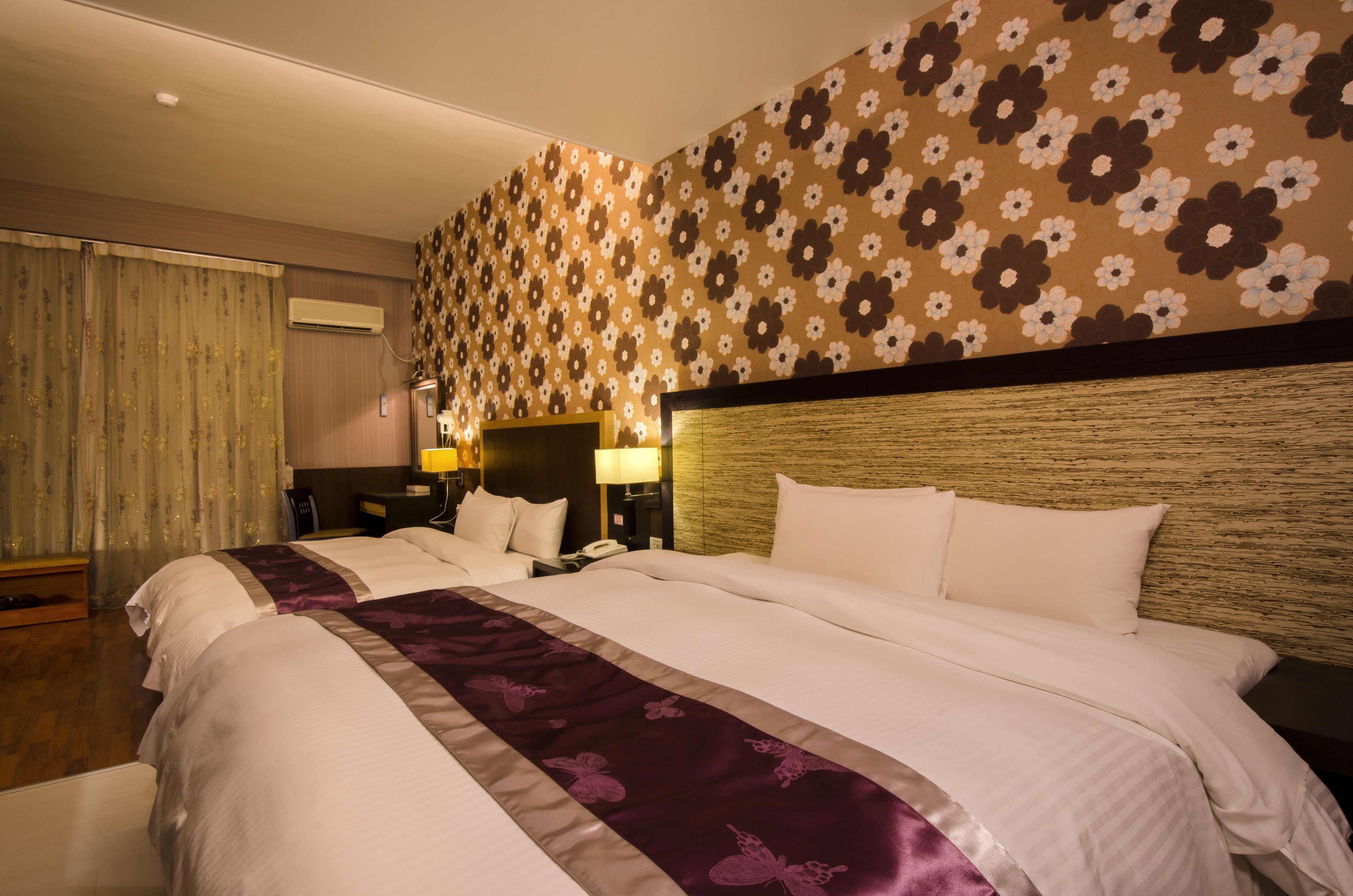 A hotel room with a large bed, a nightstand, a lamp, and a floral-patterned wall.