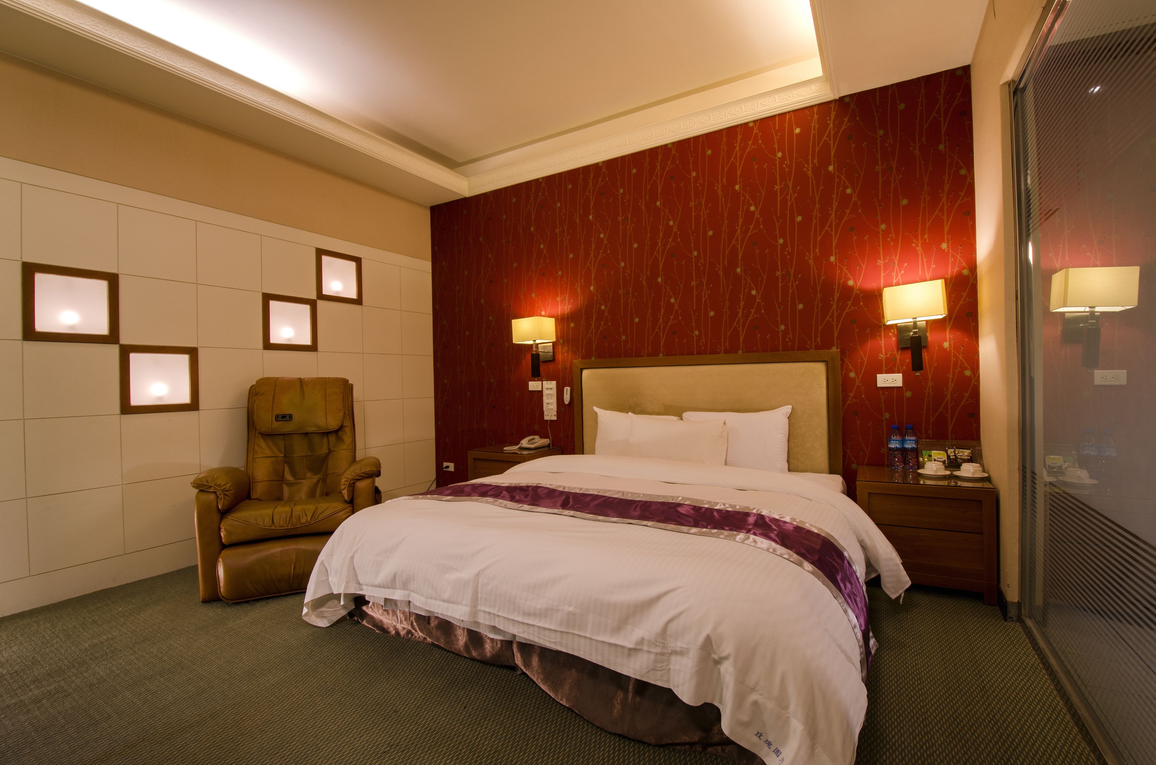 Habitación de hotel con una cama grande, un sillón de masajes, dos lámparas de noche y una mesita de noche con un teléfono y botellas de agua.