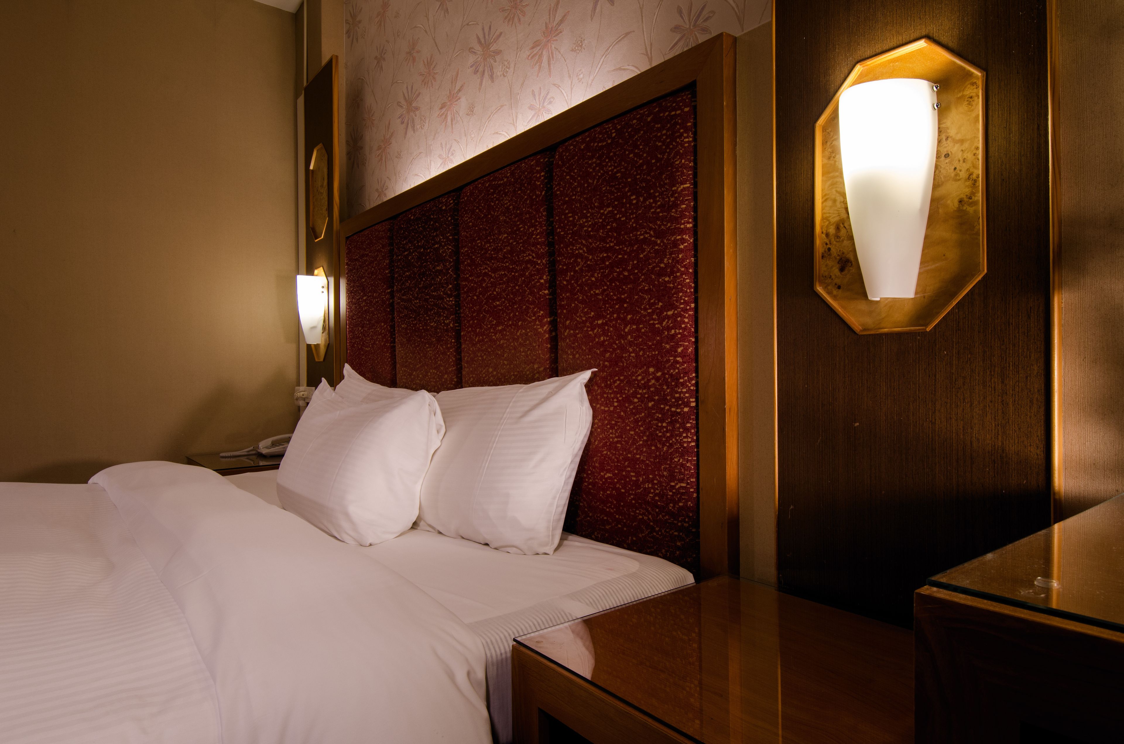 A hotel room with a large bed, a wooden headboard, a nightstand with a lamp, and a wall-mounted light fixture.
