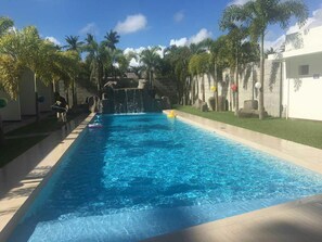 Piscina com água azul cristalina, rodeada por palmeiras e uma cascata de pedra.