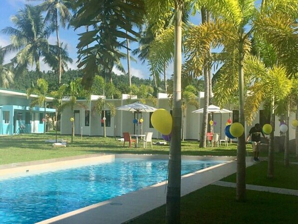 Uma área de piscina com piscina azul translúcida, cercada por palmeiras e alguns edifícios de design moderno e simples.