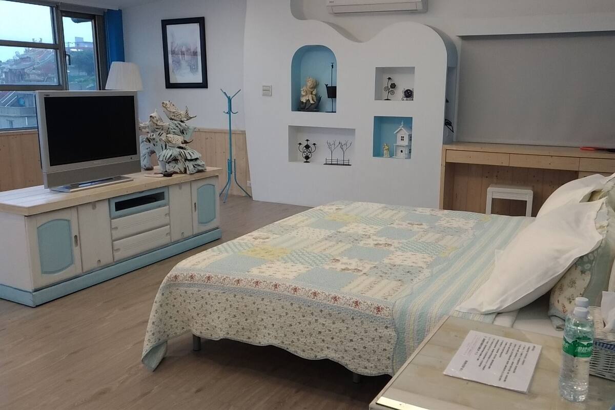Ein Schlafzimmer mit einem Bett, einem Fernseher auf einem Schrank, einem Schreibtisch mit Stuhl und einem Fenster mit Blick auf Gebäude.