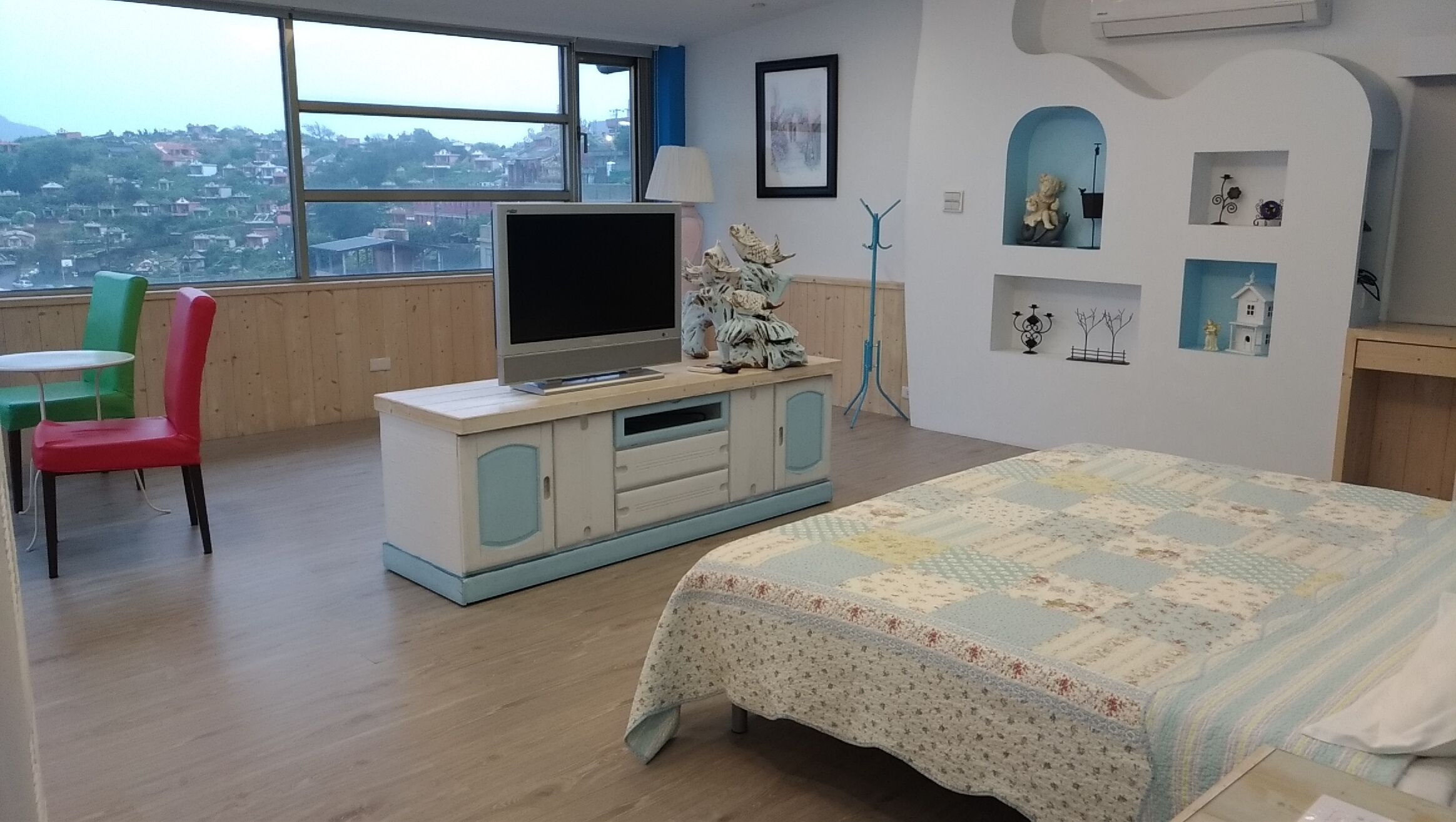 Ein Schlafzimmer mit einem Bett, einem Fernseher auf einem Schrank, einem Sessel und Blick auf eine Stadtkulisse durch ein großes Fenster.