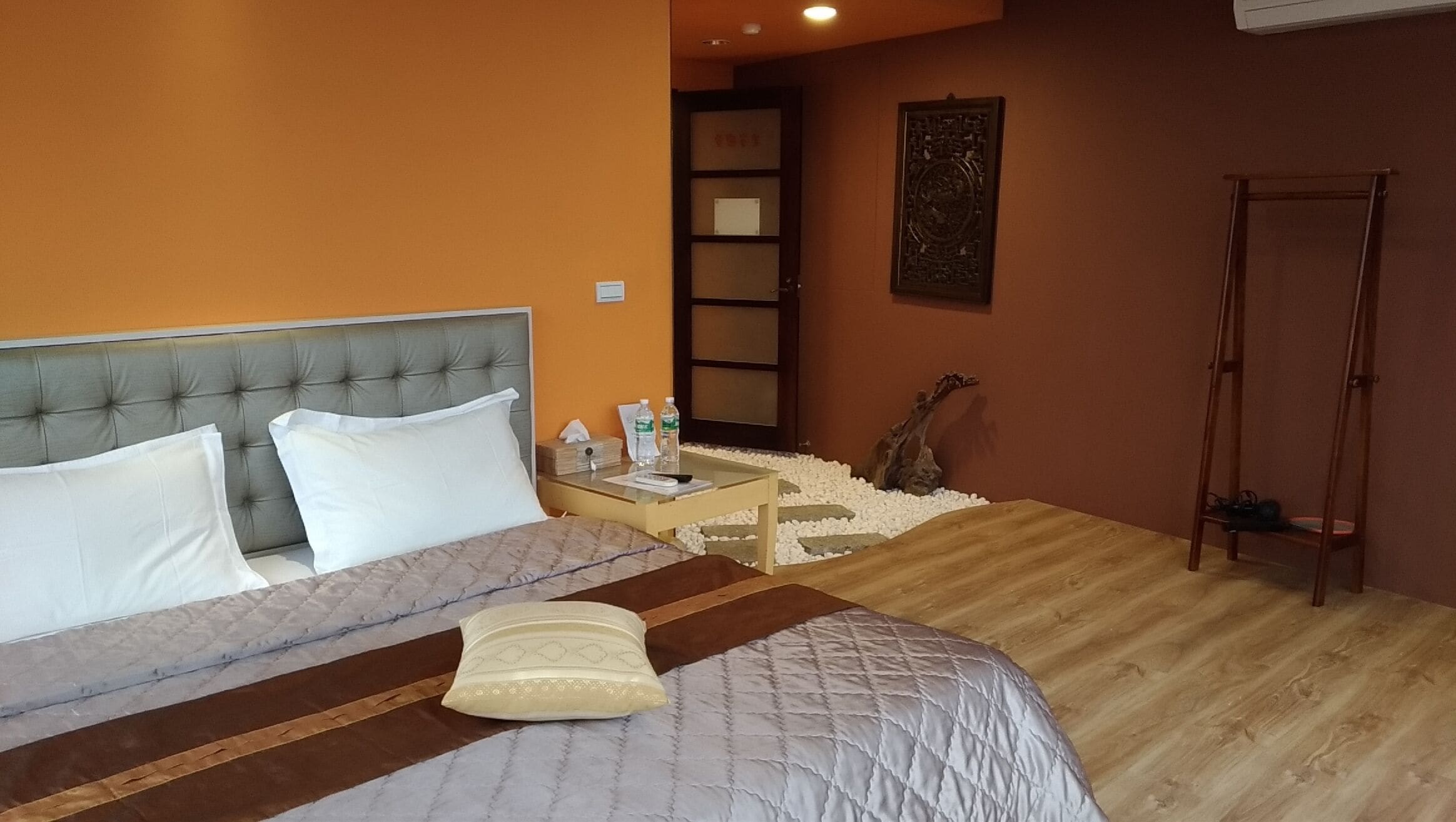 A bedroom with a bed, a nightstand, a wooden floor, a door, and a wall-mounted artwork.