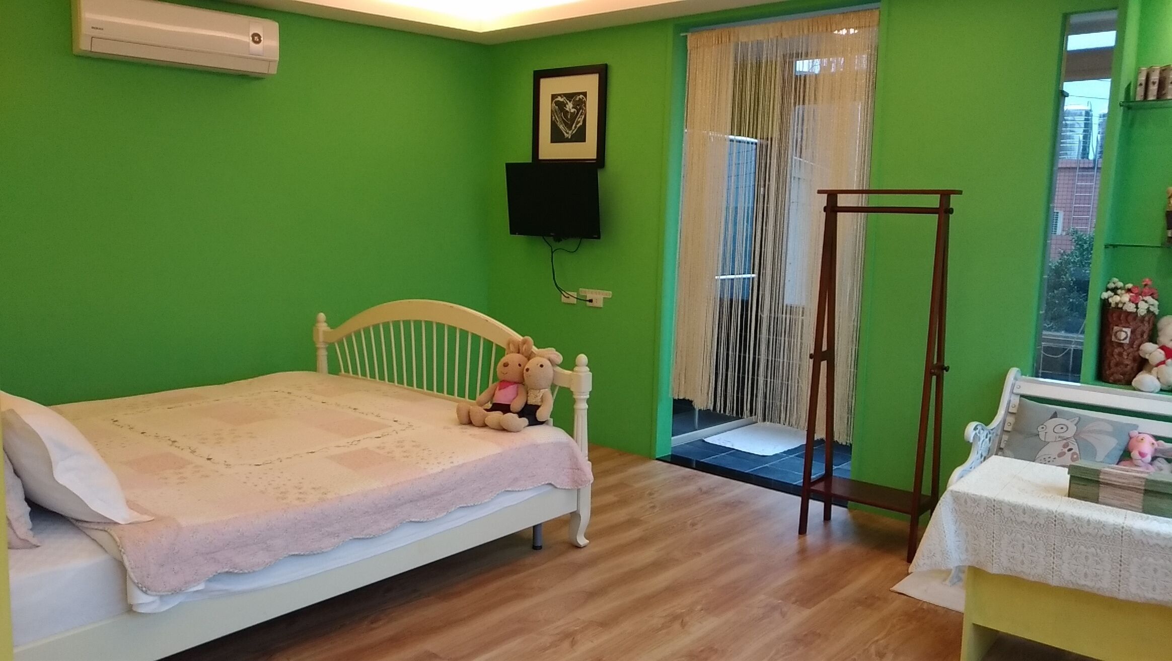 A bedroom with a bed, a wooden chair, a small table with a lamp, a window with curtains, and a wall-mounted TV.