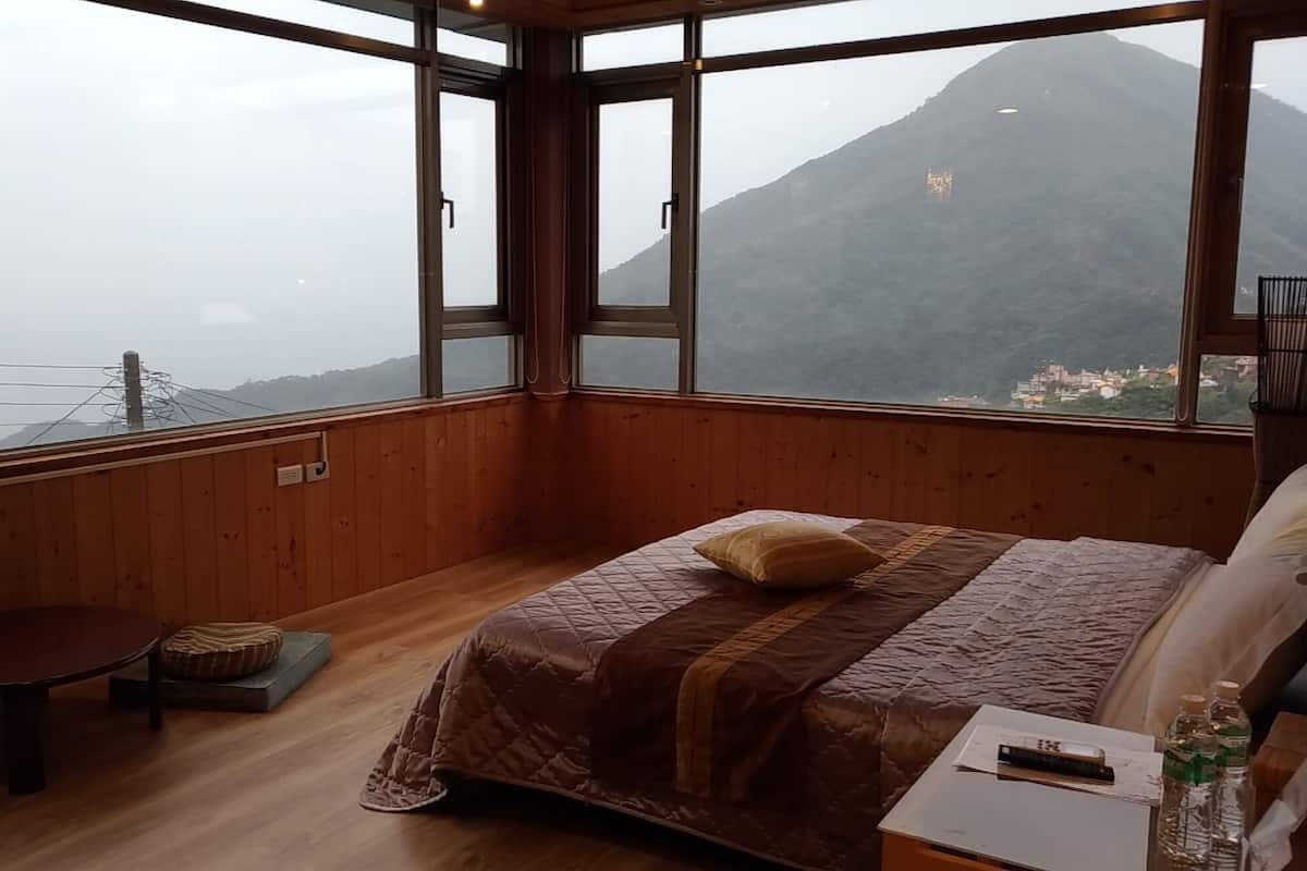 Ein Schlafzimmer mit großem Fenster, Holzwänden, einem Bett mit Tagesdecke, einem Nachttisch und einem Sessel.