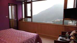 A bedroom with a bed, a wooden dresser, a TV, and a window with a view of a mountain. - Good Time B&B (New Taipei City)