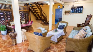A spacious living area with wicker furniture, a staircase, and a TV mounted on the wall. - Banchan Hostel (Ayutthaya)