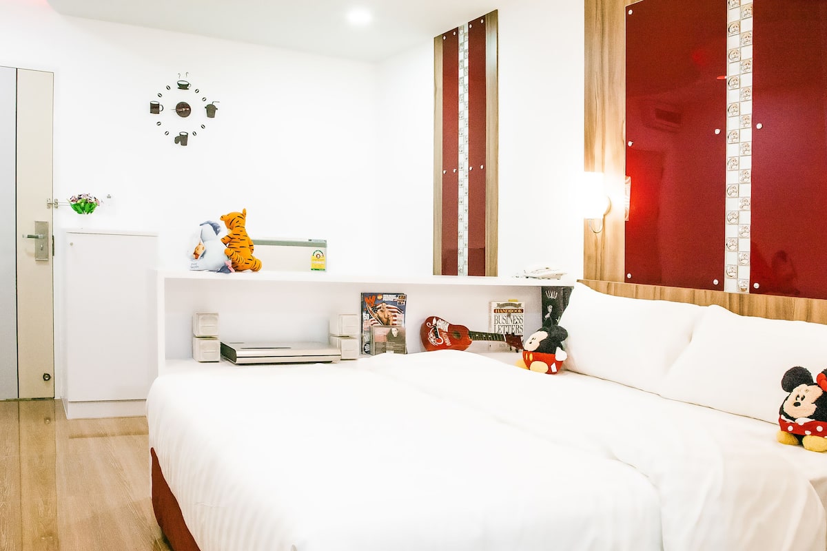 A bedroom with a bed, a desk, a wall clock, and a window with curtains.