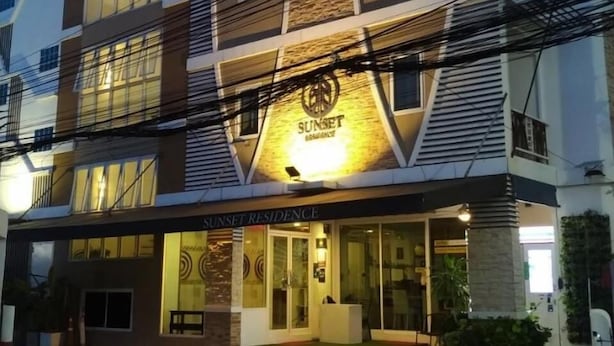A multi-story building with a prominent logo on the facade, a ground-level entrance, and a parking area in front. - Sunset Residence Pinklao (Bangkok)