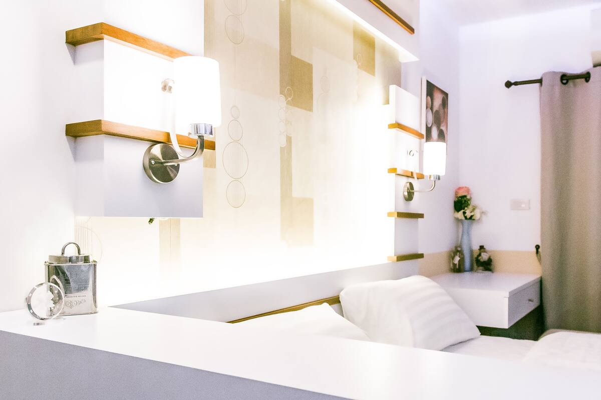 A modern hotel room with a white bed, a bedside table, a lamp, and a mirror.