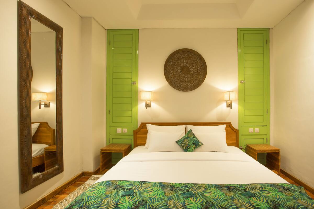 A bedroom with a bed, green shutters, a round wall decoration, bedside lamps, and a mirror.