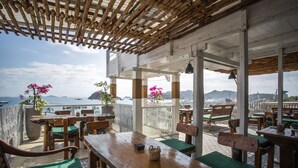 An outdoor patio with wooden tables and chairs, potted plants, and a view of the sea. - Eco Tree O'tel (Labuan Bajo)