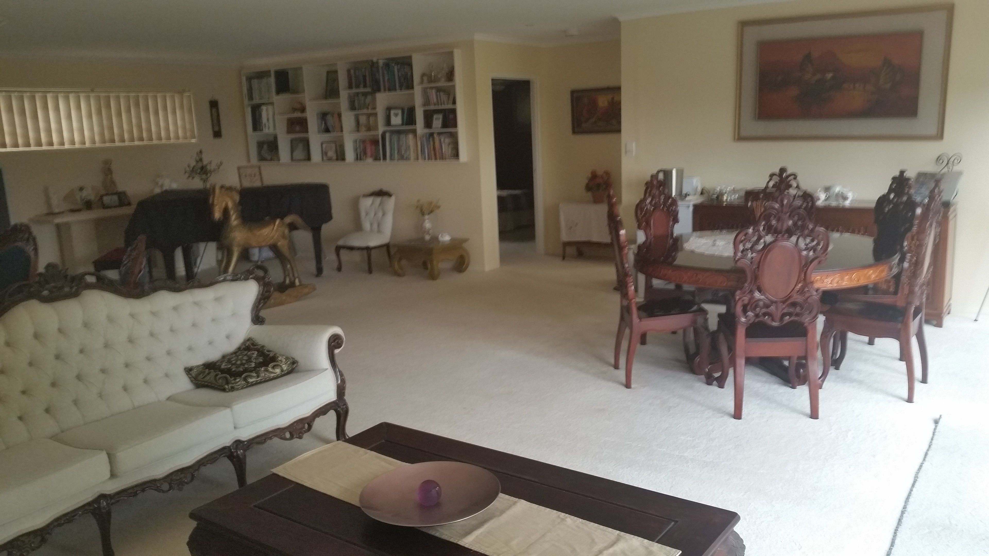 A living room with a white sofa, a wooden coffee table, a dining table with ornate chairs, a bookshelf, and a painting on the wall.