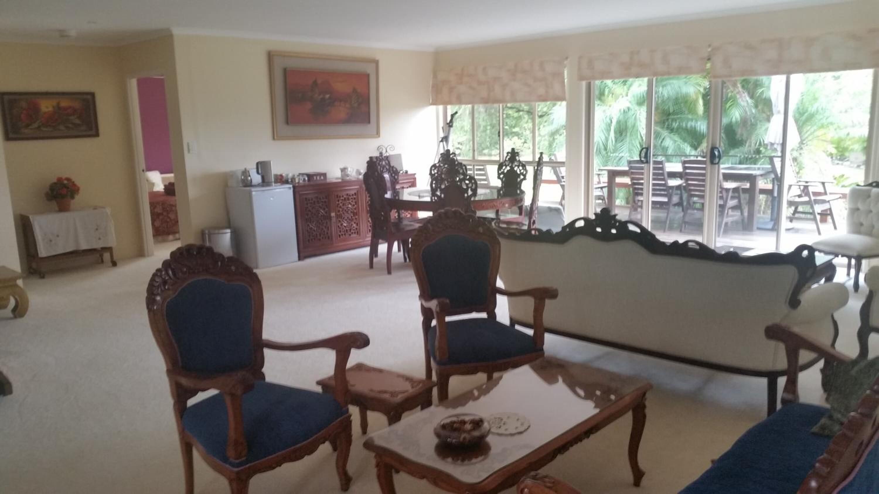 Una sala de estar con dos sillones de madera, una mesa de centro, una mesa de comedor con sillas, un refrigerador y una ventana con vista a palmeras.