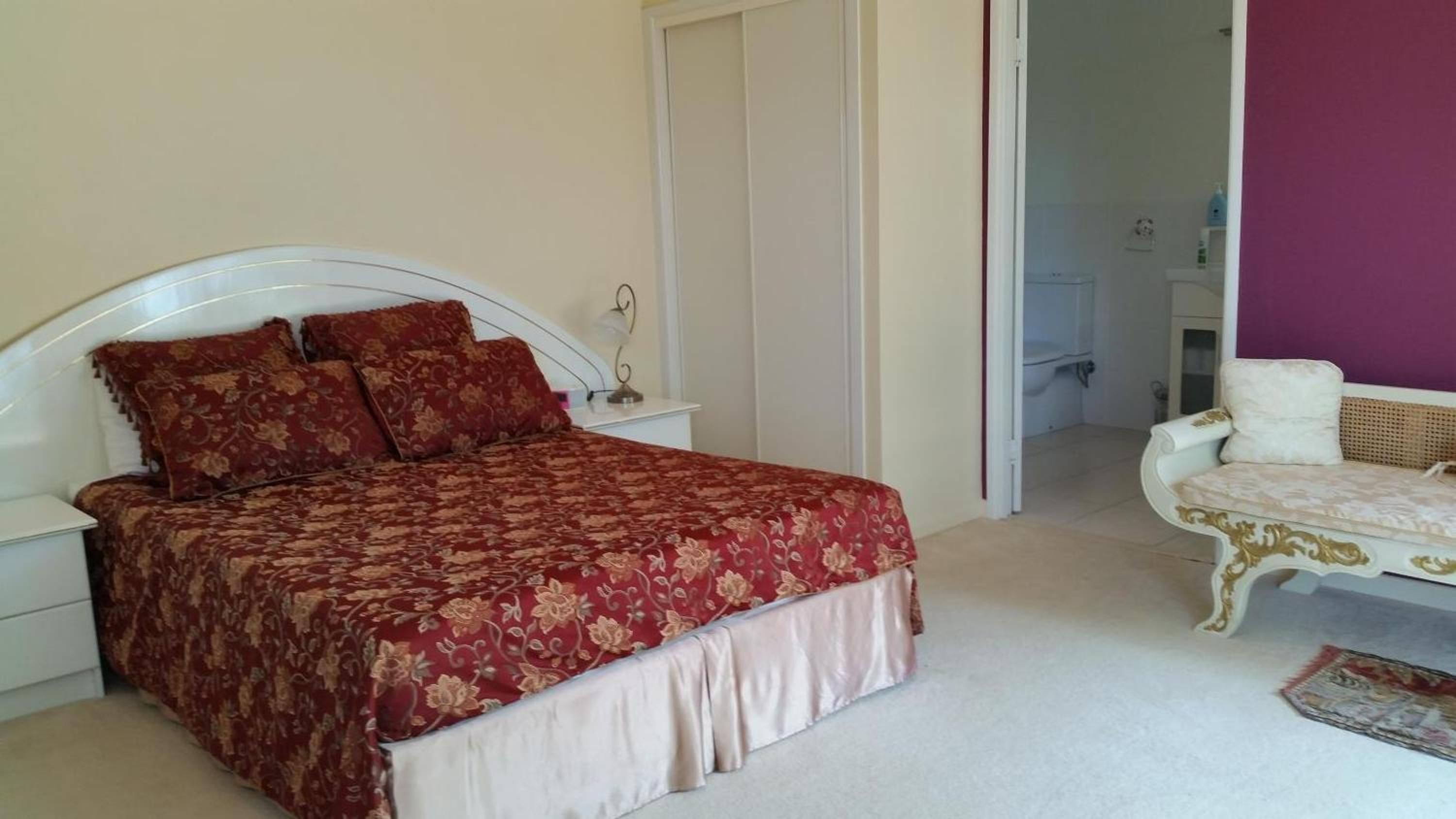 A bedroom with a large bed, a nightstand, a dresser, a chair, and a bathroom visible through an open door.