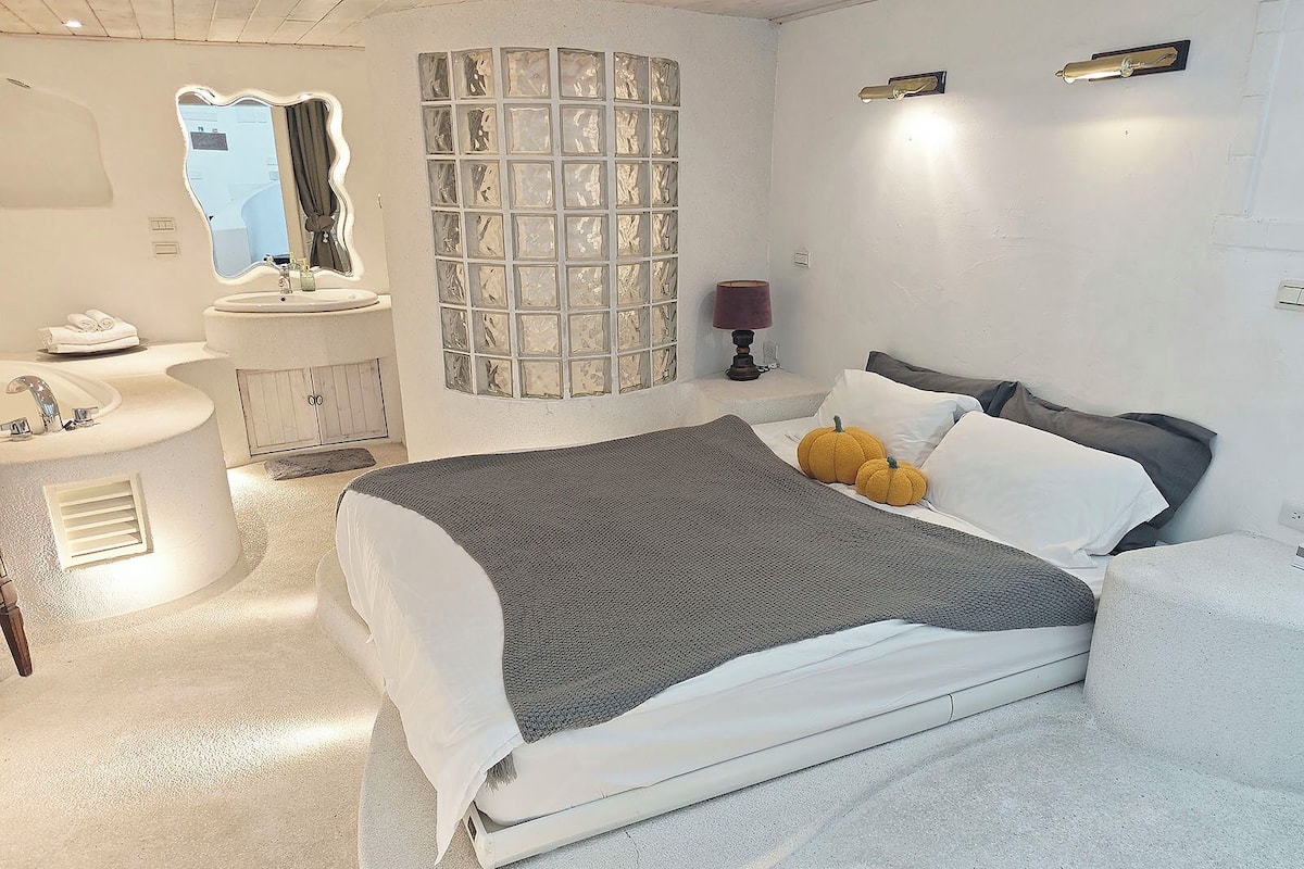 A bedroom with a bed, pillows, a nightstand, a mirror, a window with a view, and a bathroom with a sink and mirror.