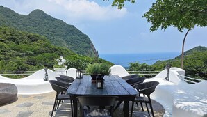 A wooden table with wicker chairs, surrounded by white walls, overlooking a mountainous landscape and the sea. - Gold Stone Crescent Moon Light (New Taipei City)