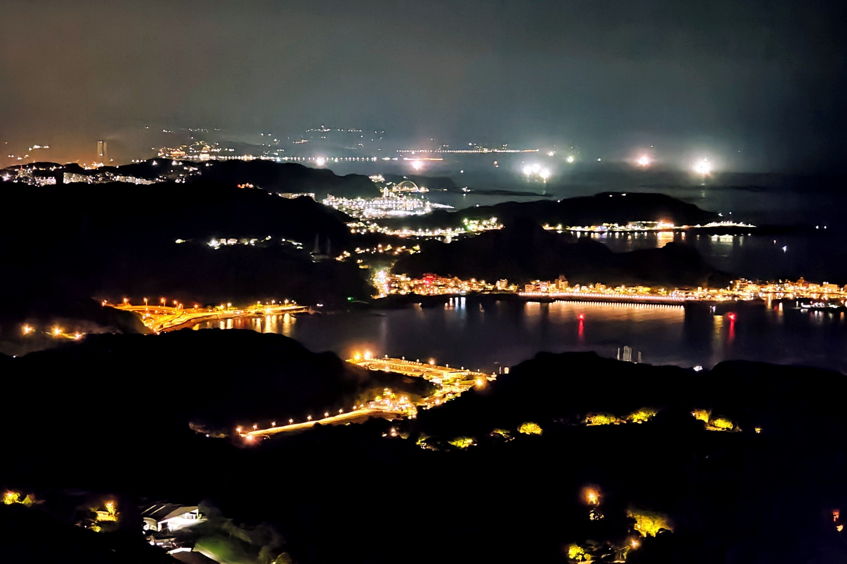 A nighttime view of a coastal city with illuminated buildings and a bridge over water.