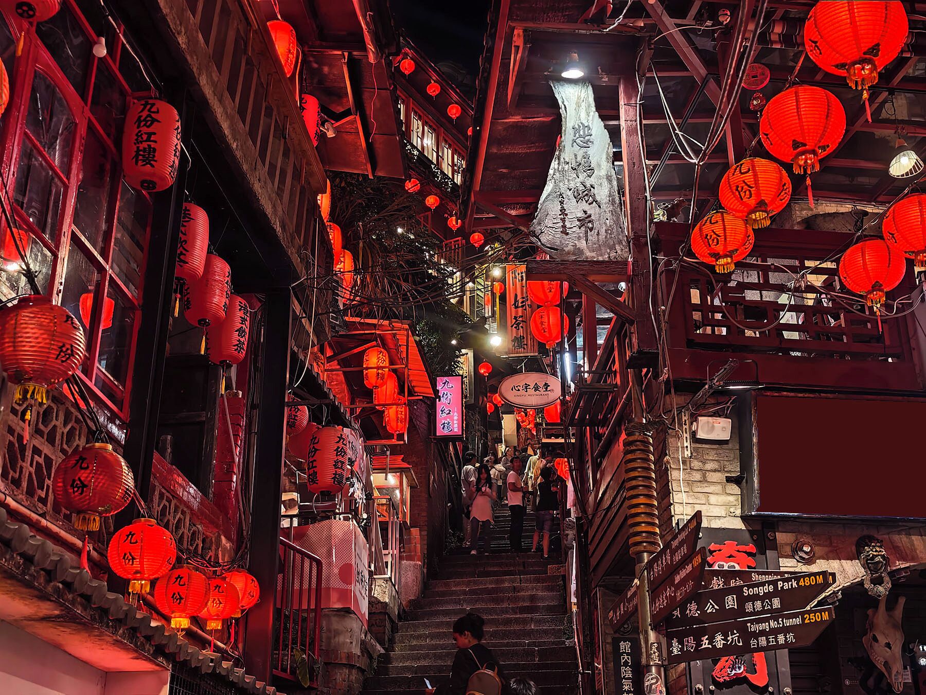 A narrow alleyway with red lanterns hanging overhead and signs in a foreign script.