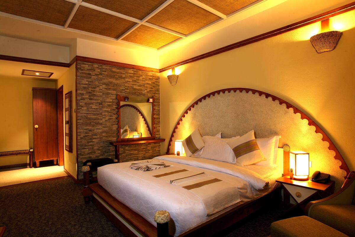 A hotel room with a large bed, a wooden headboard, a nightstand with a lamp, and a stone accent wall.