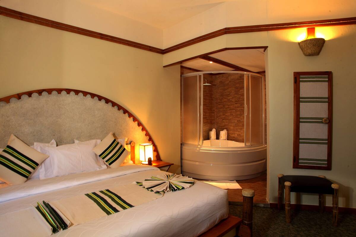 A hotel room with a large bed, a shower, a bench, and a wall-mounted shelf.