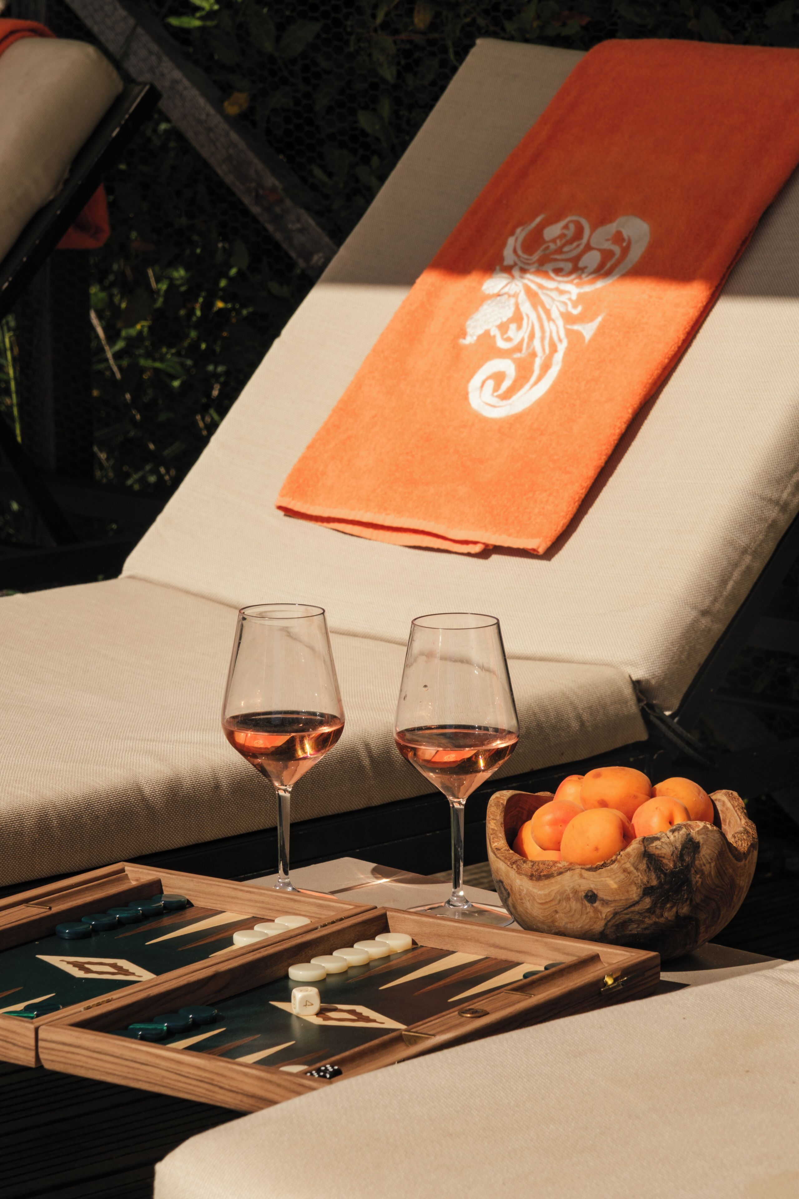 A sun lounger with a towel, two glasses of wine, a wooden board game, and a bowl of oranges.