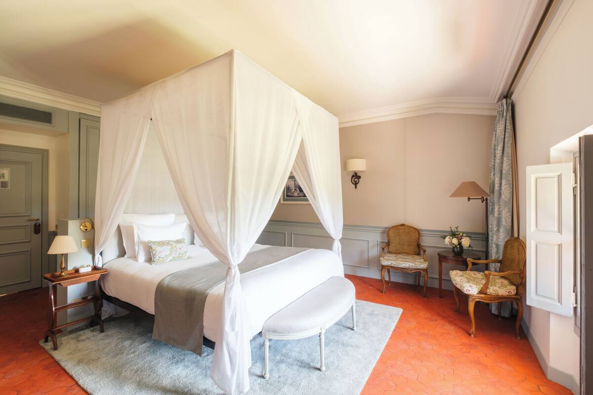 A four-poster bed with a canopy, a bedside table with a lamp, a chair, and a small table with flowers.