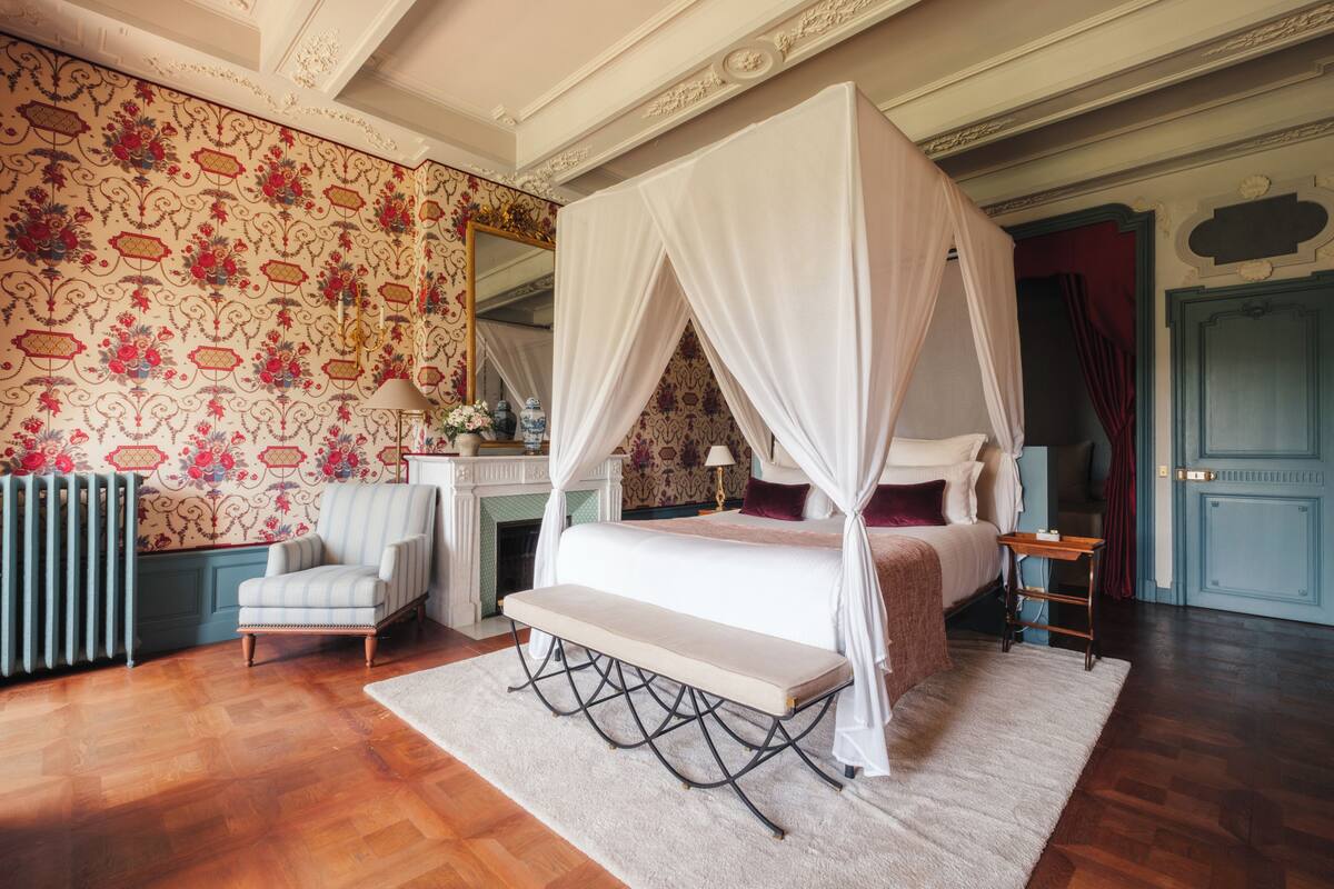 A bedroom with a four-poster bed, a bench, a chair, a mirror, a fireplace, and a radiator.
