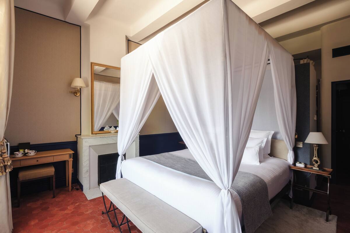 A bedroom with a four-poster bed, a bedside table, a bench, a mirror, and a fireplace.