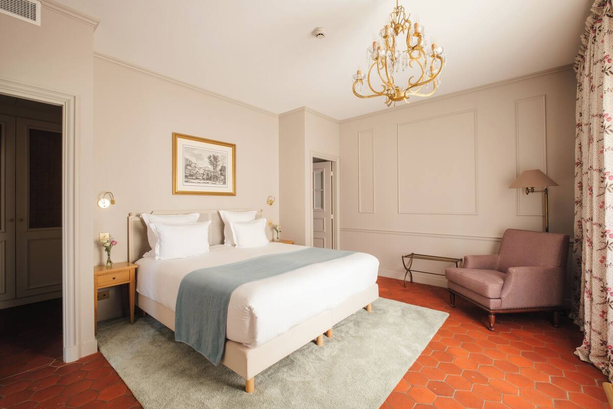 A bedroom with a bed, a chair, a chandelier, and a framed picture on the wall.