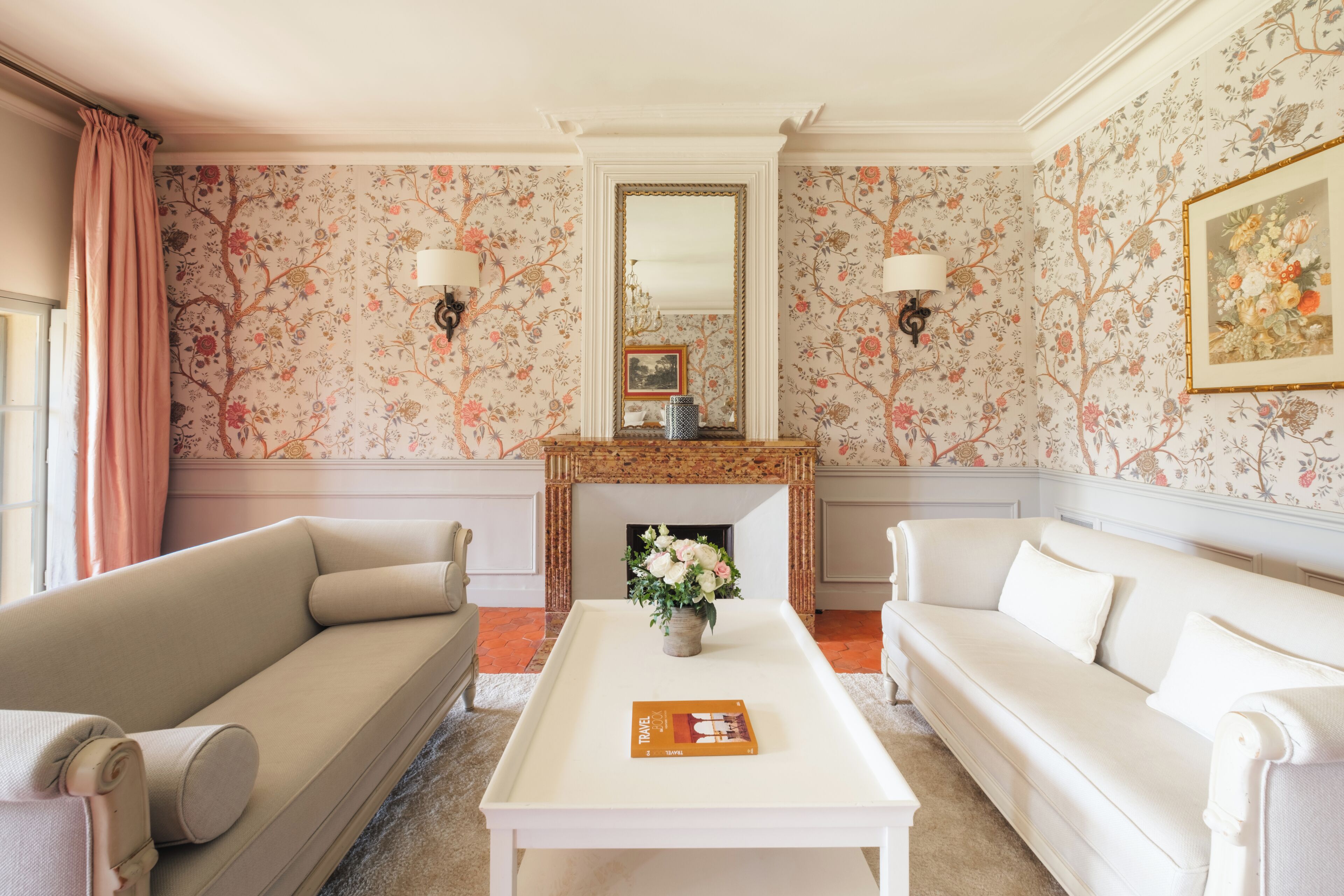 Un salon avec un papier peint à motifs floraux, une cheminée, deux canapés et une table basse.