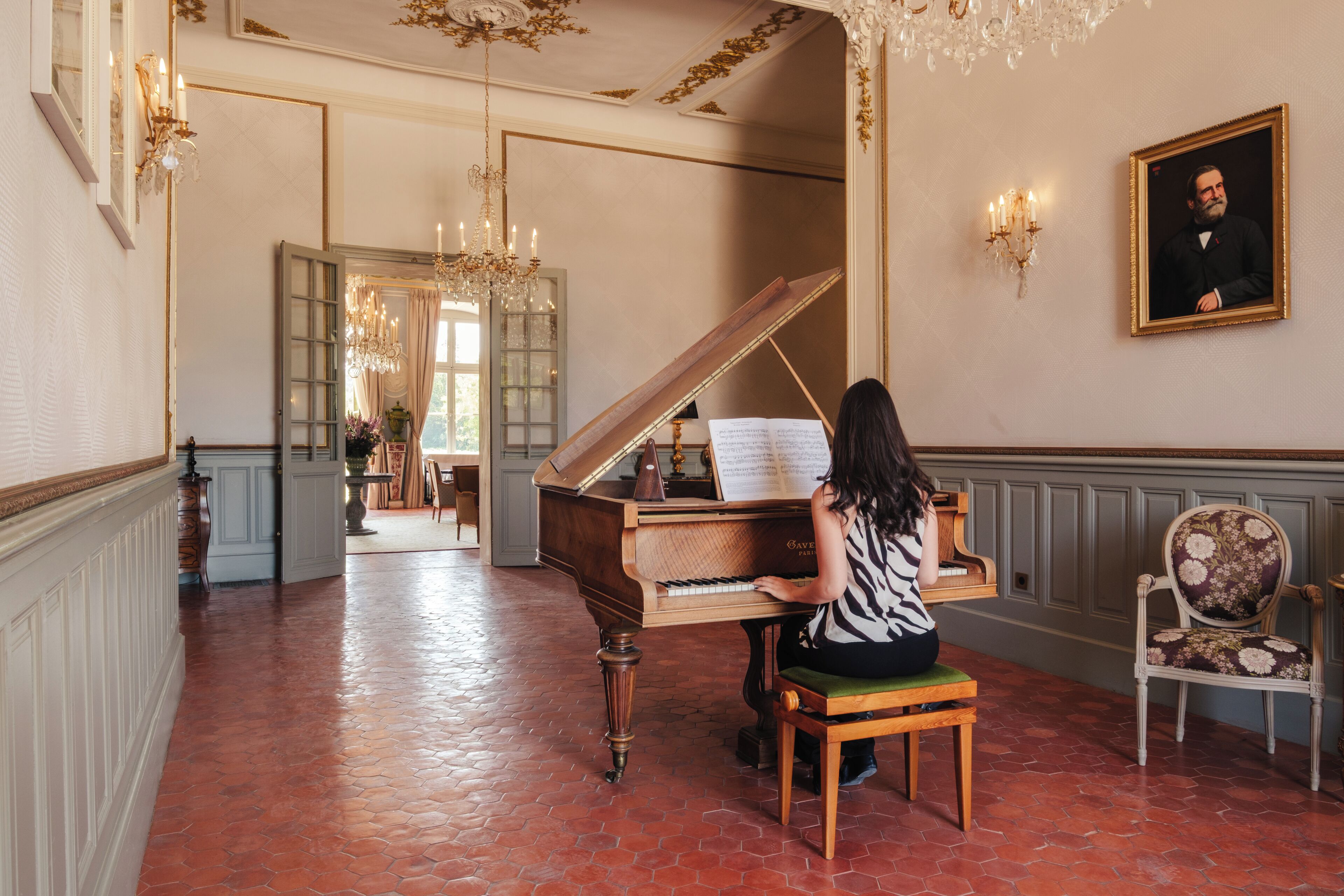 A grand piano in a spacious room with a chandelier, a framed portrait on the wall, and a chair.