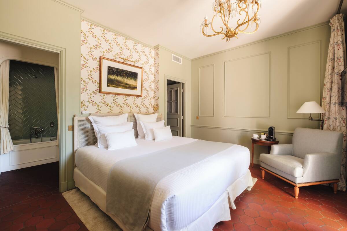 A hotel room with a large bed, a chair, a side table, and a chandelier.