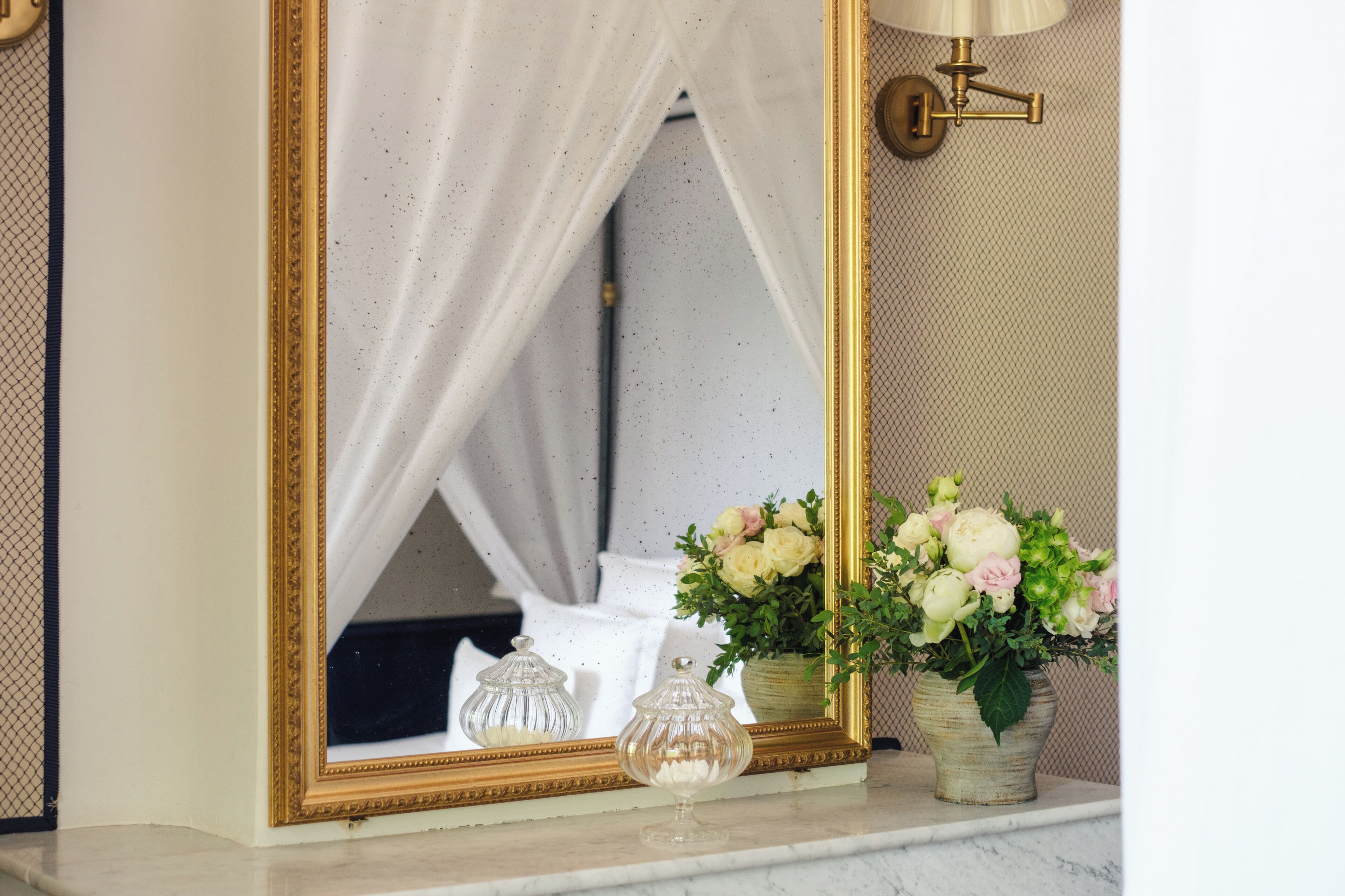 Un miroir avec un cadre doré, un dais blanc et deux vases en verre ornés de fleurs, le tout posé sur une surface en marbre.