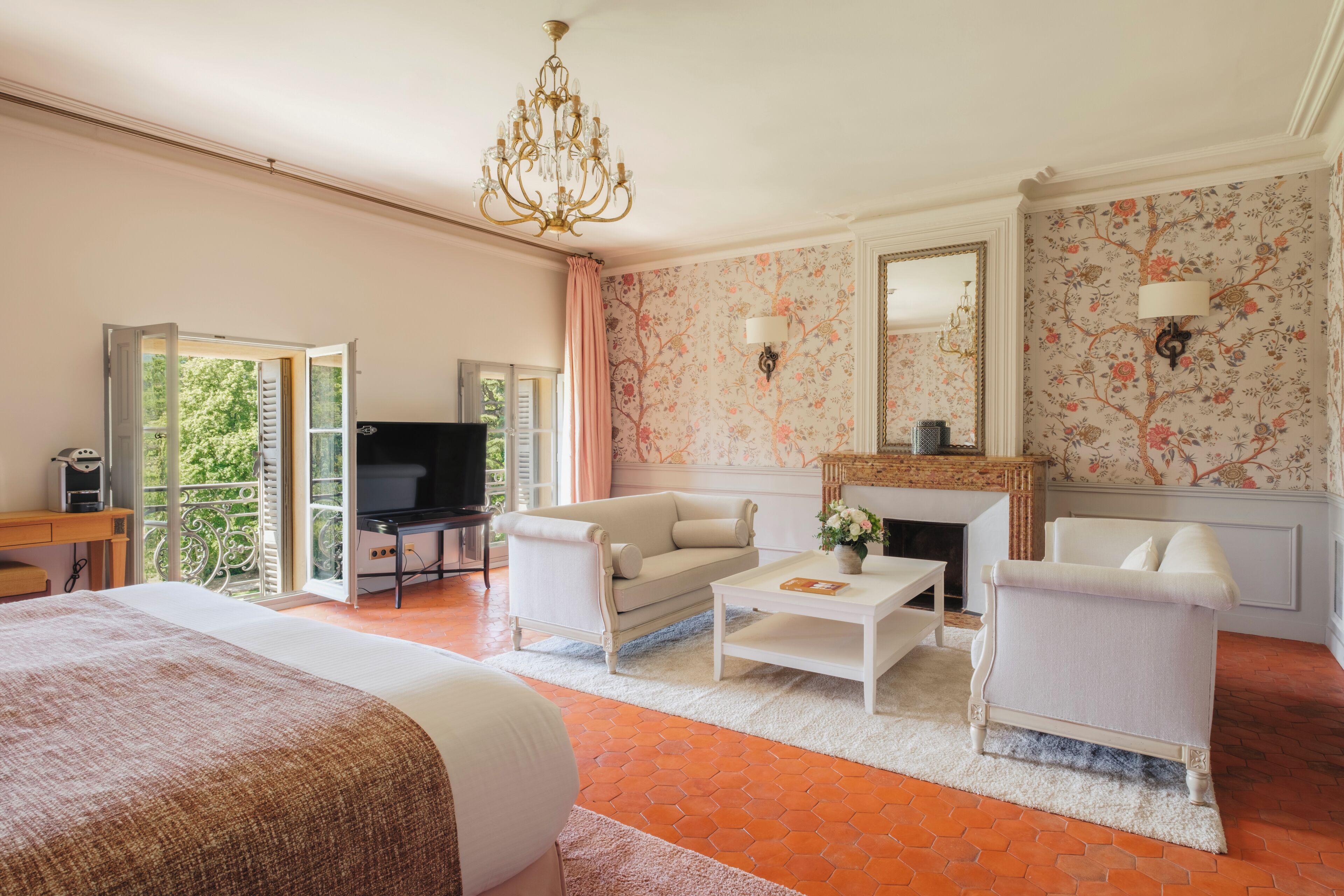 Une chambre d’hôtel avec un lit, deux fauteuils blancs, une table basse, un téléviseur, une cheminée et un balcon avec vue.