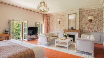 Une chambre d’hôtel avec un lit, deux fauteuils blancs, une table basse, un téléviseur, une cheminée et un balcon avec vue.